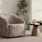 A modern tub-style accent armchair featuring a curved continuous barrel backrest, textured woven upholstery pattern, and a fully upholstered low-profile base set in a sunlit contemporary room layout.
