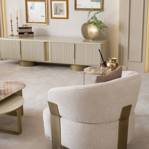 A luxurious modular Grandeur sofa set in a bright, modern living room. The set features cream-colored fabric upholstery with rectangular cushions and sleek gold metallic legs. Beside the modular sections sits a matching rounded armchair with gold accents. The scene includes minimalist decor, a marble coffee table, and large windows, highlighting the elegant design of the Grandeur furniture from Turkey.