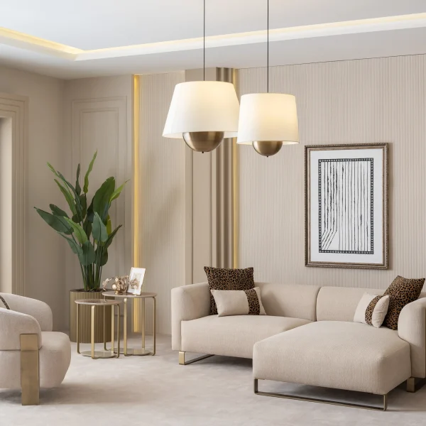 A luxurious modular Grandeur sofa set in a bright, modern living room. The set features cream-colored fabric upholstery with rectangular cushions and sleek gold metallic legs. Beside the modular sections sits a matching rounded armchair with gold accents. The scene includes minimalist decor, a marble coffee table, and large windows, highlighting the elegant design of the Grandeur furniture from Turkey.