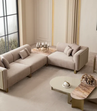A luxurious modular Grandeur sofa set in a bright, modern living room. The set features cream-colored fabric upholstery with rectangular cushions and sleek gold metallic legs. Beside the modular sections sits a matching rounded armchair with gold accents. The scene includes minimalist decor, a marble coffee table, and large windows, highlighting the elegant design of the Grandeur furniture from Turkey.