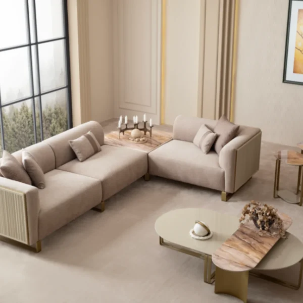 A luxurious modular Grandeur sofa set in a bright, modern living room. The set features cream-colored fabric upholstery with rectangular cushions and sleek gold metallic legs. Beside the modular sections sits a matching rounded armchair with gold accents. The scene includes minimalist decor, a marble coffee table, and large windows, highlighting the elegant design of the Grandeur furniture from Turkey.