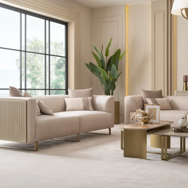 A luxurious modular Grandeur sofa set in a bright, modern living room. The set features cream-colored fabric upholstery with rectangular cushions and sleek gold metallic legs. Beside the modular sections sits a matching rounded armchair with gold accents. The scene includes minimalist decor, a marble coffee table, and large windows, highlighting the elegant design of the Grandeur furniture from Turkey.