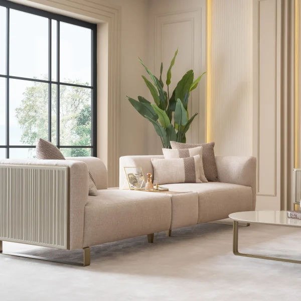 A luxurious modular Grandeur sofa set in a bright, modern living room. The set features cream-colored fabric upholstery with rectangular cushions and sleek gold metallic legs. Beside the modular sections sits a matching rounded armchair with gold accents. The scene includes minimalist decor, a marble coffee table, and large windows, highlighting the elegant design of the Grandeur furniture from Turkey.