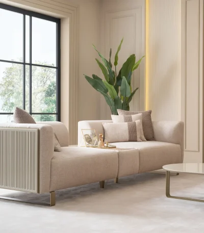 A luxurious modular Grandeur sofa set in a bright, modern living room. The set features cream-colored fabric upholstery with rectangular cushions and sleek gold metallic legs. Beside the modular sections sits a matching rounded armchair with gold accents. The scene includes minimalist decor, a marble coffee table, and large windows, highlighting the elegant design of the Grandeur furniture from Turkey.