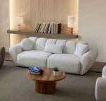 Alure Sofa Set featuring a crisp white three-seater sofa, a soft sage green loveseat, and a matching white armchair. The pieces showcase deep vertical stitching and a plush, rounded design, centered around a fluted wooden coffee table in a minimalist, warm-toned living room.