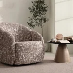 A modern tub-style accent armchair featuring a curved continuous barrel backrest, textured woven upholstery pattern, and a fully upholstered low-profile base set in a sunlit contemporary room layout.
