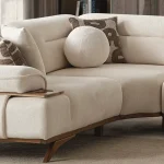 A modern living room featuring the Grace Sofa. The arrangement displays a plush taupe sofa and a matching cream sofa, both designed with distinctive integrated wooden side tables on the armrests and angled wooden legs. A white swivel armchair and a round wooden coffee table complete the cozy, contemporary look of this furniture from Turkey.