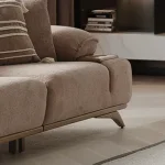 A modern living room featuring the Grace Sofa. The arrangement displays a plush taupe sofa and a matching cream sofa, both designed with distinctive integrated wooden side tables on the armrests and angled wooden legs. A white swivel armchair and a round wooden coffee table complete the cozy, contemporary look of this furniture from Turkey.