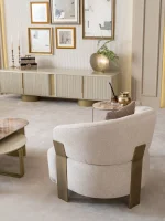 A luxurious modular Grandeur sofa set in a bright, modern living room. The set features cream-colored fabric upholstery with rectangular cushions and sleek gold metallic legs. Beside the modular sections sits a matching rounded armchair with gold accents. The scene includes minimalist decor, a marble coffee table, and large windows, highlighting the elegant design of the Grandeur furniture from Turkey.