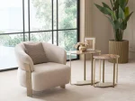 A luxurious modular Grandeur sofa set in a bright, modern living room. The set features cream-colored fabric upholstery with rectangular cushions and sleek gold metallic legs. Beside the modular sections sits a matching rounded armchair with gold accents. The scene includes minimalist decor, a marble coffee table, and large windows, highlighting the elegant design of the Grandeur furniture from Turkey.
