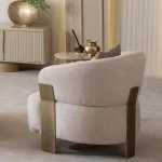 A luxurious modular Grandeur sofa set in a bright, modern living room. The set features cream-colored fabric upholstery with rectangular cushions and sleek gold metallic legs. Beside the modular sections sits a matching rounded armchair with gold accents. The scene includes minimalist decor, a marble coffee table, and large windows, highlighting the elegant design of the Grandeur furniture from Turkey.
