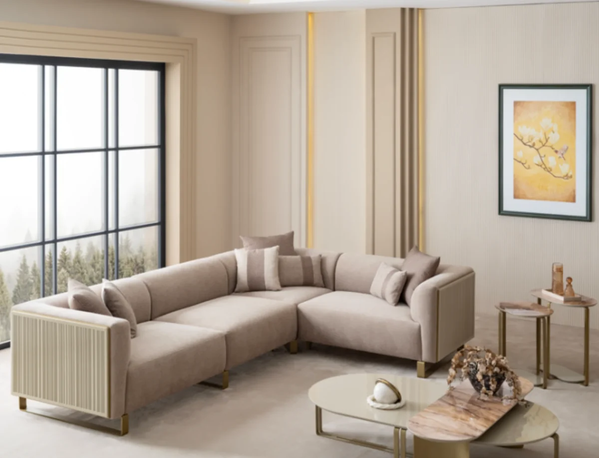 A luxurious modular Grandeur sofa set in a bright, modern living room. The set features cream-colored fabric upholstery with rectangular cushions and sleek gold metallic legs. Beside the modular sections sits a matching rounded armchair with gold accents. The scene includes minimalist decor, a marble coffee table, and large windows, highlighting the elegant design of the Grandeur furniture from Turkey.