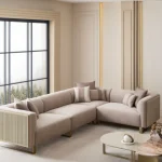 A luxurious modular Grandeur sofa set in a bright, modern living room. The set features cream-colored fabric upholstery with rectangular cushions and sleek gold metallic legs. Beside the modular sections sits a matching rounded armchair with gold accents. The scene includes minimalist decor, a marble coffee table, and large windows, highlighting the elegant design of the Grandeur furniture from Turkey.