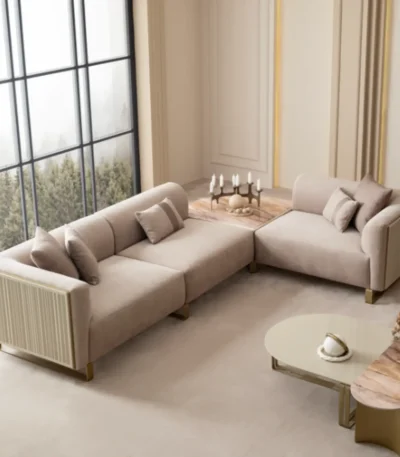A luxurious modular Grandeur sofa set in a bright, modern living room. The set features cream-colored fabric upholstery with rectangular cushions and sleek gold metallic legs. Beside the modular sections sits a matching rounded armchair with gold accents. The scene includes minimalist decor, a marble coffee table, and large windows, highlighting the elegant design of the Grandeur furniture from Turkey.