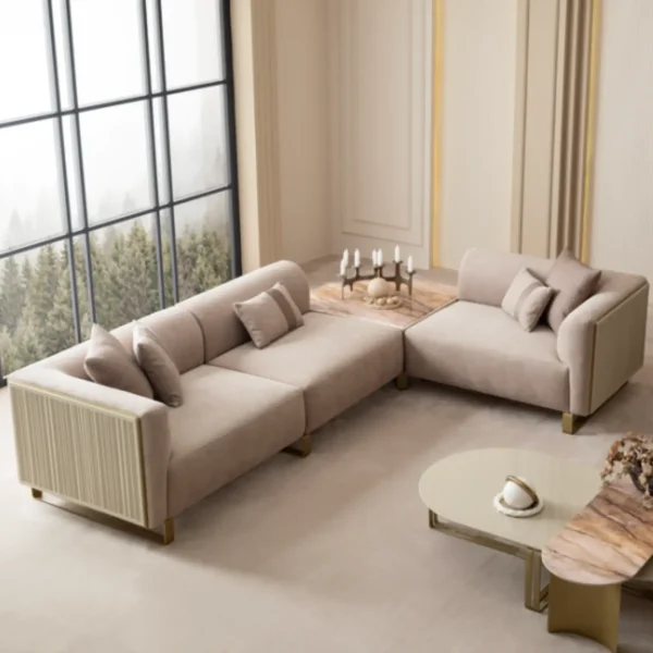 A luxurious modular Grandeur sofa set in a bright, modern living room. The set features cream-colored fabric upholstery with rectangular cushions and sleek gold metallic legs. Beside the modular sections sits a matching rounded armchair with gold accents. The scene includes minimalist decor, a marble coffee table, and large windows, highlighting the elegant design of the Grandeur furniture from Turkey.