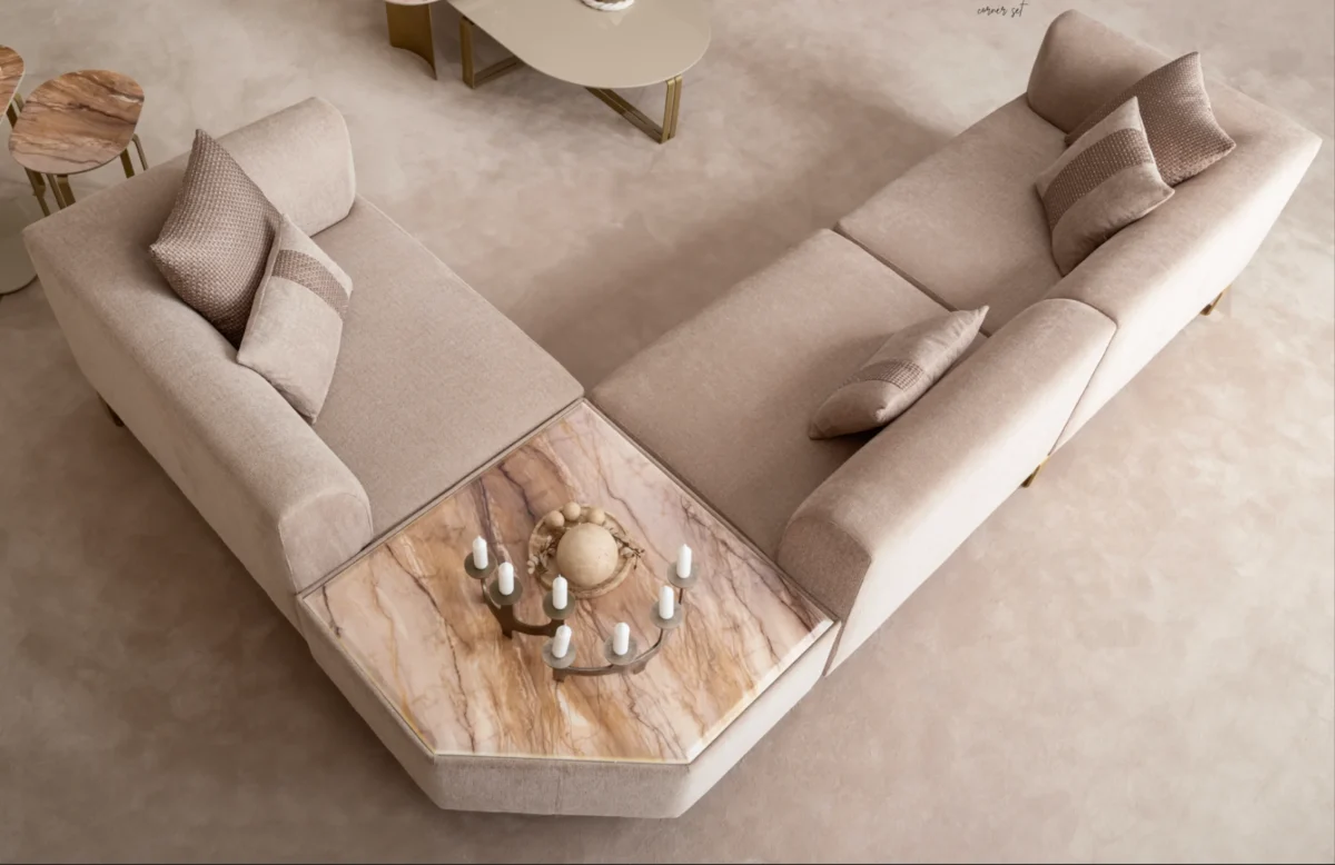 A luxurious modular Grandeur sofa set in a bright, modern living room. The set features cream-colored fabric upholstery with rectangular cushions and sleek gold metallic legs. Beside the modular sections sits a matching rounded armchair with gold accents. The scene includes minimalist decor, a marble coffee table, and large windows, highlighting the elegant design of the Grandeur furniture from Turkey.