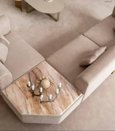 A luxurious modular Grandeur sofa set in a bright, modern living room. The set features cream-colored fabric upholstery with rectangular cushions and sleek gold metallic legs. Beside the modular sections sits a matching rounded armchair with gold accents. The scene includes minimalist decor, a marble coffee table, and large windows, highlighting the elegant design of the Grandeur furniture from Turkey.