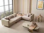 A luxurious modular Grandeur sofa set in a bright, modern living room. The set features cream-colored fabric upholstery with rectangular cushions and sleek gold metallic legs. Beside the modular sections sits a matching rounded armchair with gold accents. The scene includes minimalist decor, a marble coffee table, and large windows, highlighting the elegant design of the Grandeur furniture from Turkey.