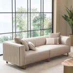 A luxurious modular Grandeur sofa set in a bright, modern living room. The set features cream-colored fabric upholstery with rectangular cushions and sleek gold metallic legs. Beside the modular sections sits a matching rounded armchair with gold accents. The scene includes minimalist decor, a marble coffee table, and large windows, highlighting the elegant design of the Grandeur furniture from Turkey.