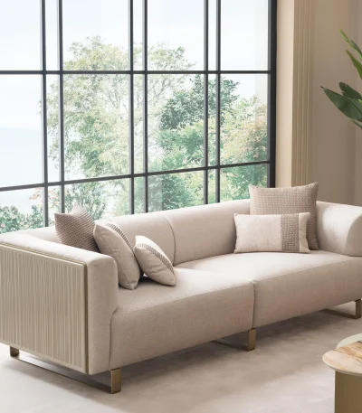 Luksuzna modularna garnitura sofa Grandeur u svetloj, modernoj dnevnoj sobi. Garnitura je presvučena kremastom tkaninom sa pravougaonim jastucima i elegantnim zlatnim metalnim nogama. Pored modularnih delova nalazi se odgovarajuća okrugla fotelja sa zlatnim akcentima. Scena uključuje minimalistički dekor, mermerni stočić za kafu i velike prozore, ističući elegantan dizajn nameštaja Grandeur iz Turske.
