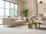 A luxurious modular Grandeur sofa set in a bright, modern living room. The set features cream-colored fabric upholstery with rectangular cushions and sleek gold metallic legs. Beside the modular sections sits a matching rounded armchair with gold accents. The scene includes minimalist decor, a marble coffee table, and large windows, highlighting the elegant design of the Grandeur furniture from Turkey.
