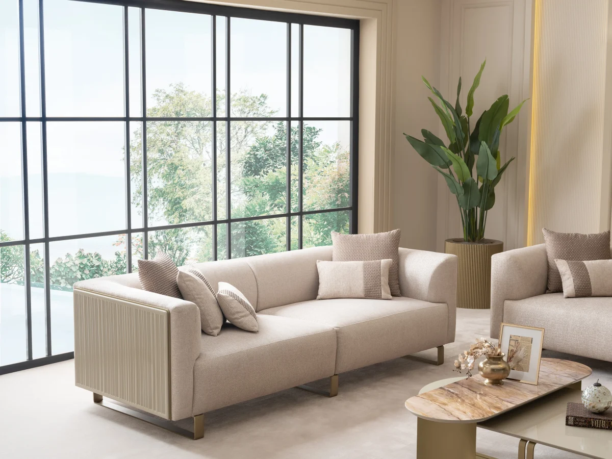 A luxurious modular Grandeur sofa set in a bright, modern living room. The set features cream-colored fabric upholstery with rectangular cushions and sleek gold metallic legs. Beside the modular sections sits a matching rounded armchair with gold accents. The scene includes minimalist decor, a marble coffee table, and large windows, highlighting the elegant design of the Grandeur furniture from Turkey.