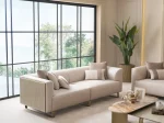 A luxurious modular Grandeur sofa set in a bright, modern living room. The set features cream-colored fabric upholstery with rectangular cushions and sleek gold metallic legs. Beside the modular sections sits a matching rounded armchair with gold accents. The scene includes minimalist decor, a marble coffee table, and large windows, highlighting the elegant design of the Grandeur furniture from Turkey.