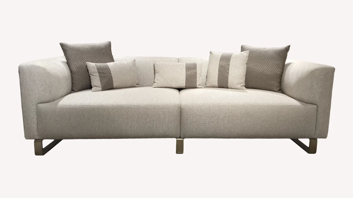 A luxurious modular Grandeur sofa set in a bright, modern living room. The set features cream-colored fabric upholstery with rectangular cushions and sleek gold metallic legs. Beside the modular sections sits a matching rounded armchair with gold accents. The scene includes minimalist decor, a marble coffee table, and large windows, highlighting the elegant design of the Grandeur furniture from Turkey.