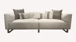 A luxurious modular Grandeur sofa set in a bright, modern living room. The set features cream-colored fabric upholstery with rectangular cushions and sleek gold metallic legs. Beside the modular sections sits a matching rounded armchair with gold accents. The scene includes minimalist decor, a marble coffee table, and large windows, highlighting the elegant design of the Grandeur furniture from Turkey.