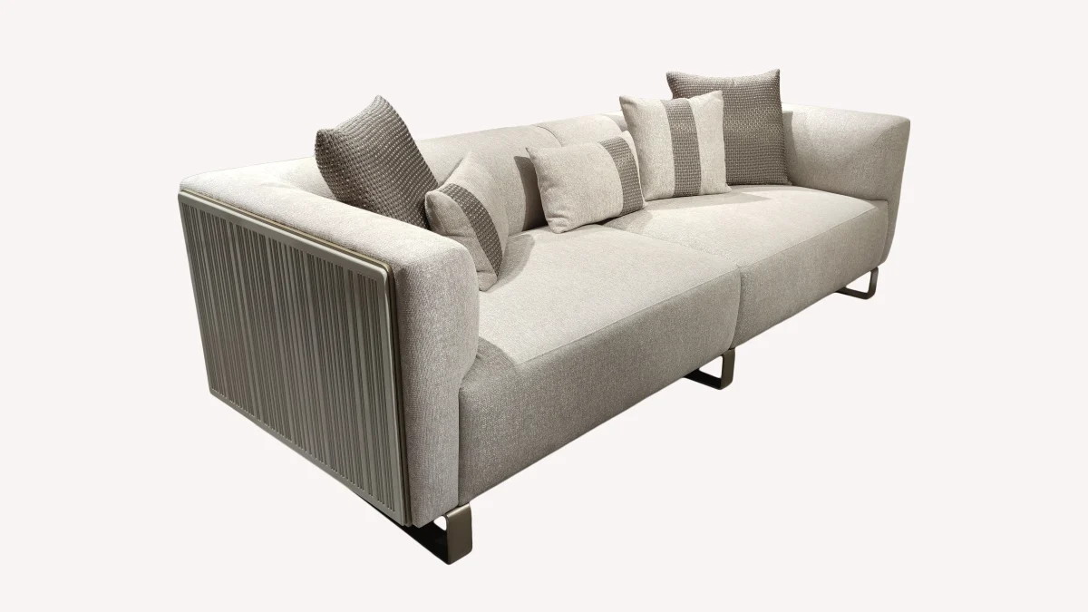 A luxurious modular Grandeur sofa set in a bright, modern living room. The set features cream-colored fabric upholstery with rectangular cushions and sleek gold metallic legs. Beside the modular sections sits a matching rounded armchair with gold accents. The scene includes minimalist decor, a marble coffee table, and large windows, highlighting the elegant design of the Grandeur furniture from Turkey.