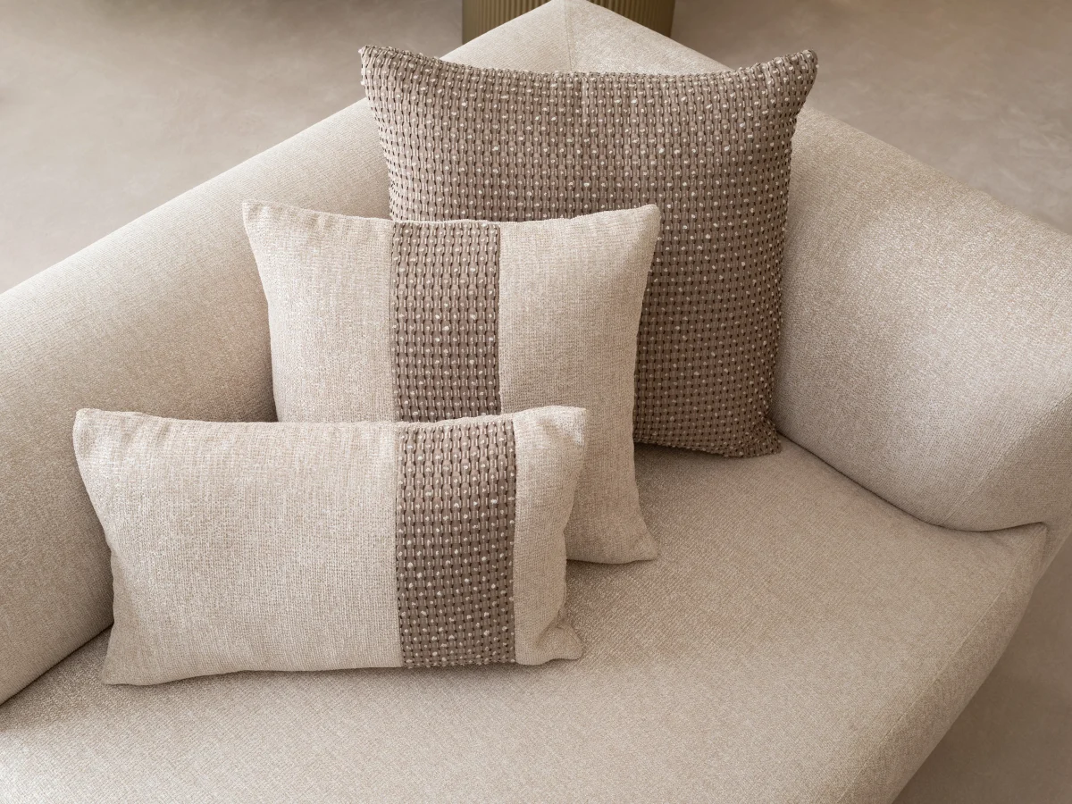 A luxurious modular Grandeur sofa set in a bright, modern living room. The set features cream-colored fabric upholstery with rectangular cushions and sleek gold metallic legs. Beside the modular sections sits a matching rounded armchair with gold accents. The scene includes minimalist decor, a marble coffee table, and large windows, highlighting the elegant design of the Grandeur furniture from Turkey.