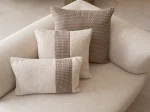 A luxurious modular Grandeur sofa set in a bright, modern living room. The set features cream-colored fabric upholstery with rectangular cushions and sleek gold metallic legs. Beside the modular sections sits a matching rounded armchair with gold accents. The scene includes minimalist decor, a marble coffee table, and large windows, highlighting the elegant design of the Grandeur furniture from Turkey.