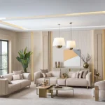 A luxurious modular Grandeur sofa set in a bright, modern living room. The set features cream-colored fabric upholstery with rectangular cushions and sleek gold metallic legs. Beside the modular sections sits a matching rounded armchair with gold accents. The scene includes minimalist decor, a marble coffee table, and large windows, highlighting the elegant design of the Grandeur furniture from Turkey.