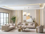 A luxurious modular Grandeur sofa set in a bright, modern living room. The set features cream-colored fabric upholstery with rectangular cushions and sleek gold metallic legs. Beside the modular sections sits a matching rounded armchair with gold accents. The scene includes minimalist decor, a marble coffee table, and large windows, highlighting the elegant design of the Grandeur furniture from Turkey.