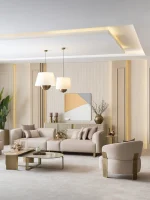 A luxurious modular Grandeur sofa set in a bright, modern living room. The set features cream-colored fabric upholstery with rectangular cushions and sleek gold metallic legs. Beside the modular sections sits a matching rounded armchair with gold accents. The scene includes minimalist decor, a marble coffee table, and large windows, highlighting the elegant design of the Grandeur furniture from Turkey.