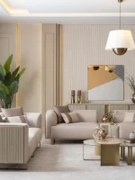A luxurious modular Grandeur sofa set in a bright, modern living room. The set features cream-colored fabric upholstery with rectangular cushions and sleek gold metallic legs. Beside the modular sections sits a matching rounded armchair with gold accents. The scene includes minimalist decor, a marble coffee table, and large windows, highlighting the elegant design of the Grandeur furniture from Turkey.