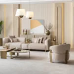A luxurious modular Grandeur sofa set in a bright, modern living room. The set features cream-colored fabric upholstery with rectangular cushions and sleek gold metallic legs. Beside the modular sections sits a matching rounded armchair with gold accents. The scene includes minimalist decor, a marble coffee table, and large windows, highlighting the elegant design of the Grandeur furniture from Turkey.