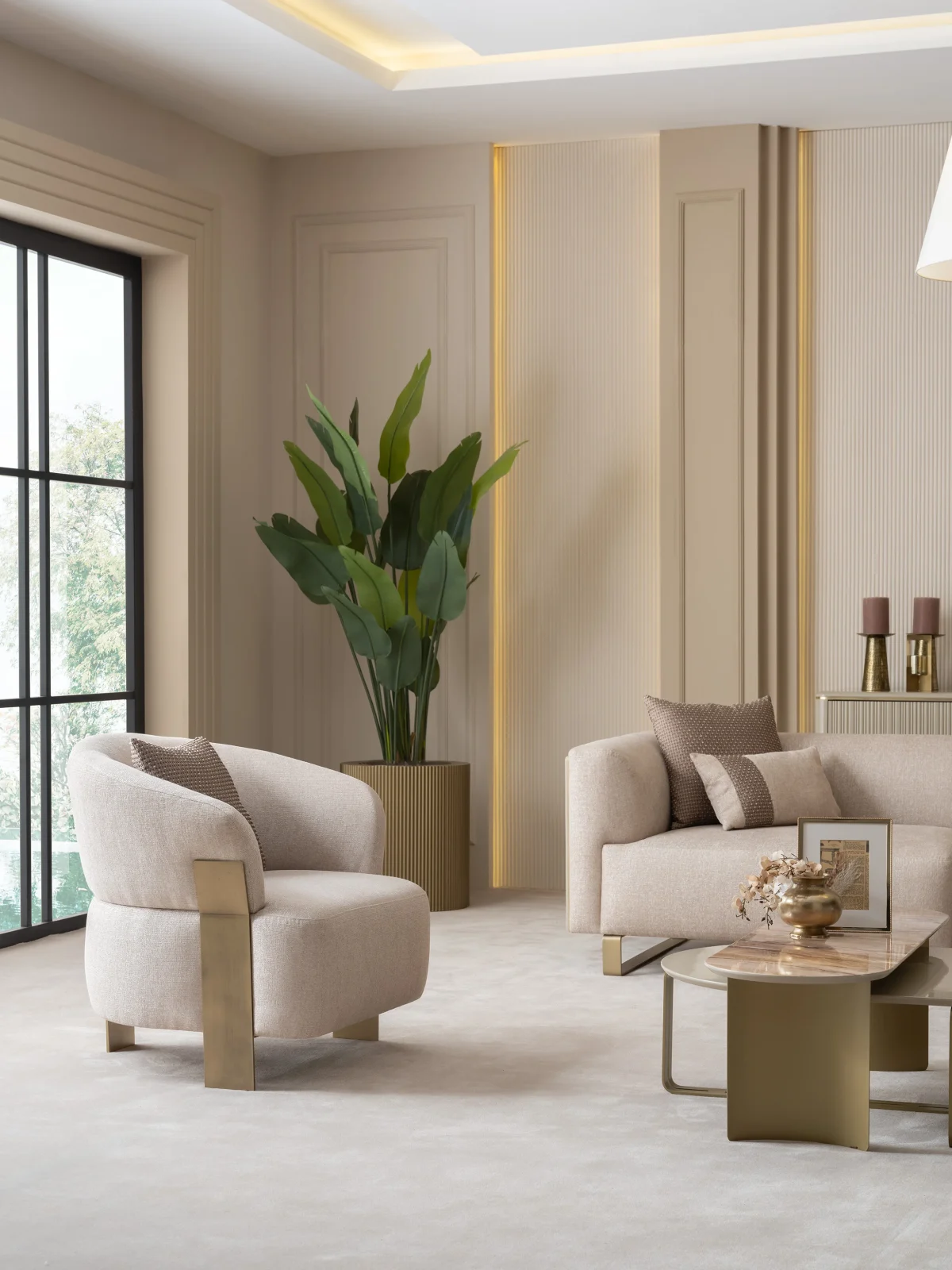 A luxurious modular Grandeur sofa set in a bright, modern living room. The set features cream-colored fabric upholstery with rectangular cushions and sleek gold metallic legs. Beside the modular sections sits a matching rounded armchair with gold accents. The scene includes minimalist decor, a marble coffee table, and large windows, highlighting the elegant design of the Grandeur furniture from Turkey.