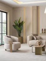 A luxurious modular Grandeur sofa set in a bright, modern living room. The set features cream-colored fabric upholstery with rectangular cushions and sleek gold metallic legs. Beside the modular sections sits a matching rounded armchair with gold accents. The scene includes minimalist decor, a marble coffee table, and large windows, highlighting the elegant design of the Grandeur furniture from Turkey.