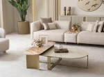 A luxurious modular Grandeur sofa set in a bright, modern living room. The set features cream-colored fabric upholstery with rectangular cushions and sleek gold metallic legs. Beside the modular sections sits a matching rounded armchair with gold accents. The scene includes minimalist decor, a marble coffee table, and large windows, highlighting the elegant design of the Grandeur furniture from Turkey.