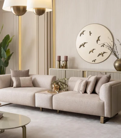Luksuzna modularna garnitura sofa Grandeur u svetloj, modernoj dnevnoj sobi. Garnitura je presvučena kremastom tkaninom sa pravougaonim jastucima i elegantnim zlatnim metalnim nogama. Pored modularnih delova nalazi se odgovarajuća okrugla fotelja sa zlatnim akcentima. Scena uključuje minimalistički dekor, mermerni stočić za kafu i velike prozore, ističući elegantan dizajn nameštaja Grandeur iz Turske.