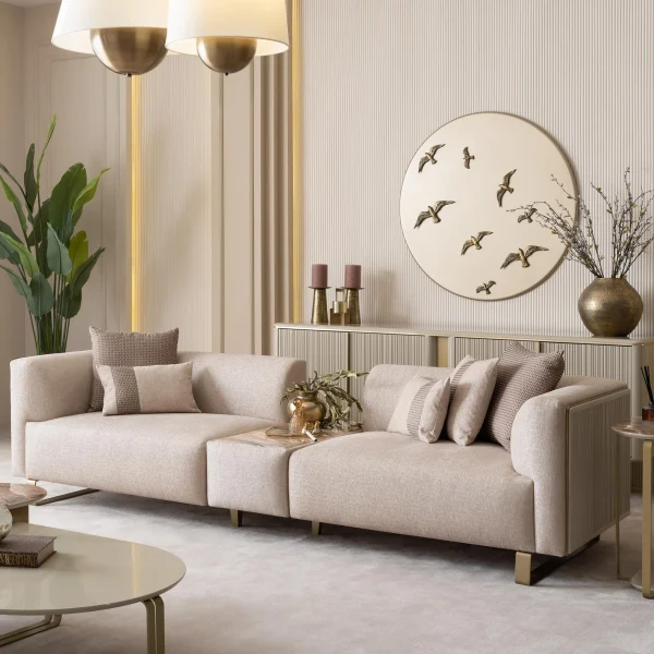A luxurious modular Grandeur sofa set in a bright, modern living room. The set features cream-colored fabric upholstery with rectangular cushions and sleek gold metallic legs. Beside the modular sections sits a matching rounded armchair with gold accents. The scene includes minimalist decor, a marble coffee table, and large windows, highlighting the elegant design of the Grandeur furniture from Turkey.