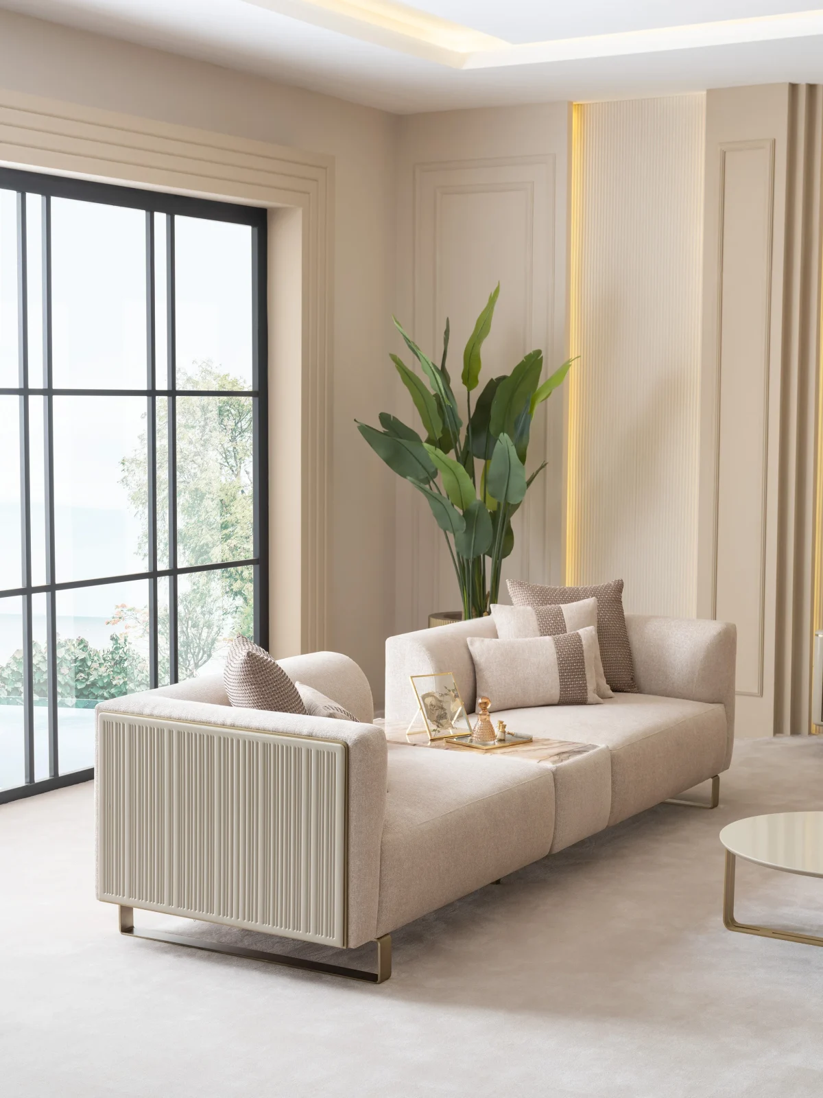 A luxurious modular Grandeur sofa set in a bright, modern living room. The set features cream-colored fabric upholstery with rectangular cushions and sleek gold metallic legs. Beside the modular sections sits a matching rounded armchair with gold accents. The scene includes minimalist decor, a marble coffee table, and large windows, highlighting the elegant design of the Grandeur furniture from Turkey.