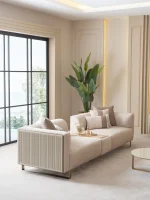 A luxurious modular Grandeur sofa set in a bright, modern living room. The set features cream-colored fabric upholstery with rectangular cushions and sleek gold metallic legs. Beside the modular sections sits a matching rounded armchair with gold accents. The scene includes minimalist decor, a marble coffee table, and large windows, highlighting the elegant design of the Grandeur furniture from Turkey.