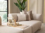 A luxurious modular Grandeur sofa set in a bright, modern living room. The set features cream-colored fabric upholstery with rectangular cushions and sleek gold metallic legs. Beside the modular sections sits a matching rounded armchair with gold accents. The scene includes minimalist decor, a marble coffee table, and large windows, highlighting the elegant design of the Grandeur furniture from Turkey.