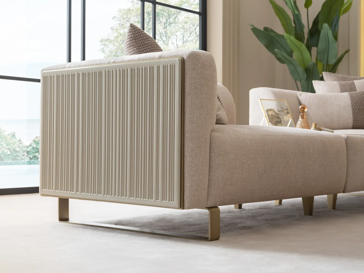 A luxurious modular Grandeur sofa set in a bright, modern living room. The set features cream-colored fabric upholstery with rectangular cushions and sleek gold metallic legs. Beside the modular sections sits a matching rounded armchair with gold accents. The scene includes minimalist decor, a marble coffee table, and large windows, highlighting the elegant design of the Grandeur furniture from Turkey.