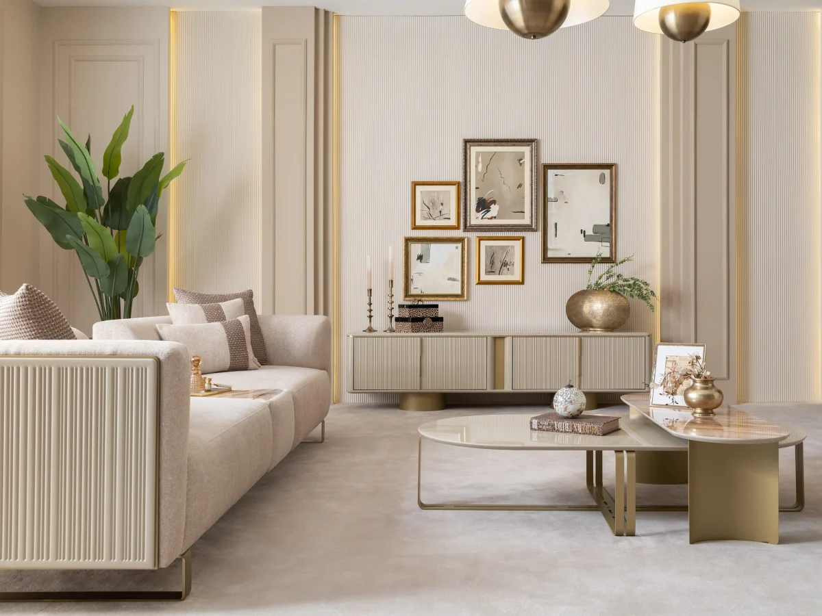 A luxurious modular Grandeur sofa set in a bright, modern living room. The set features cream-colored fabric upholstery with rectangular cushions and sleek gold metallic legs. Beside the modular sections sits a matching rounded armchair with gold accents. The scene includes minimalist decor, a marble coffee table, and large windows, highlighting the elegant design of the Grandeur furniture from Turkey.