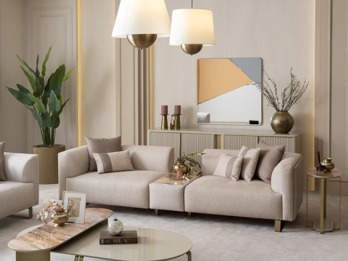 A luxurious modular Grandeur sofa set in a bright, modern living room. The set features cream-colored fabric upholstery with rectangular cushions and sleek gold metallic legs. Beside the modular sections sits a matching rounded armchair with gold accents. The scene includes minimalist decor, a marble coffee table, and large windows, highlighting the elegant design of the Grandeur furniture from Turkey.