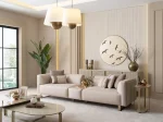 A luxurious modular Grandeur sofa set in a bright, modern living room. The set features cream-colored fabric upholstery with rectangular cushions and sleek gold metallic legs. Beside the modular sections sits a matching rounded armchair with gold accents. The scene includes minimalist decor, a marble coffee table, and large windows, highlighting the elegant design of the Grandeur furniture from Turkey.