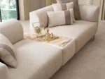 A luxurious modular Grandeur sofa set in a bright, modern living room. The set features cream-colored fabric upholstery with rectangular cushions and sleek gold metallic legs. Beside the modular sections sits a matching rounded armchair with gold accents. The scene includes minimalist decor, a marble coffee table, and large windows, highlighting the elegant design of the Grandeur furniture from Turkey.